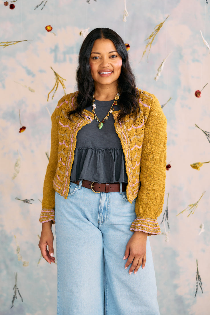 Woman wearing a mustard yellow cardigan over a dark top and light blue jeans, standing against a decorative wall.