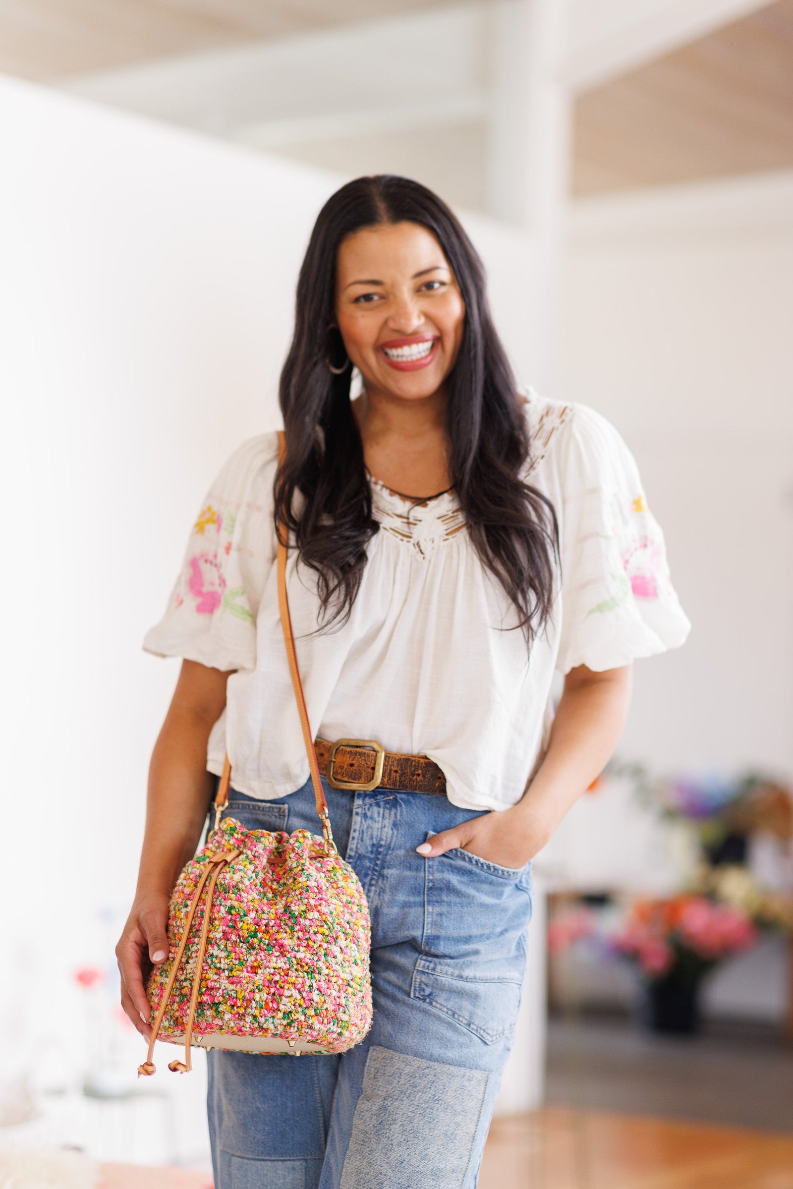 Smiling model wearing bucket bag on shoulder, resting hand toward bottom base of bag with other hand in pocket of denim jeans