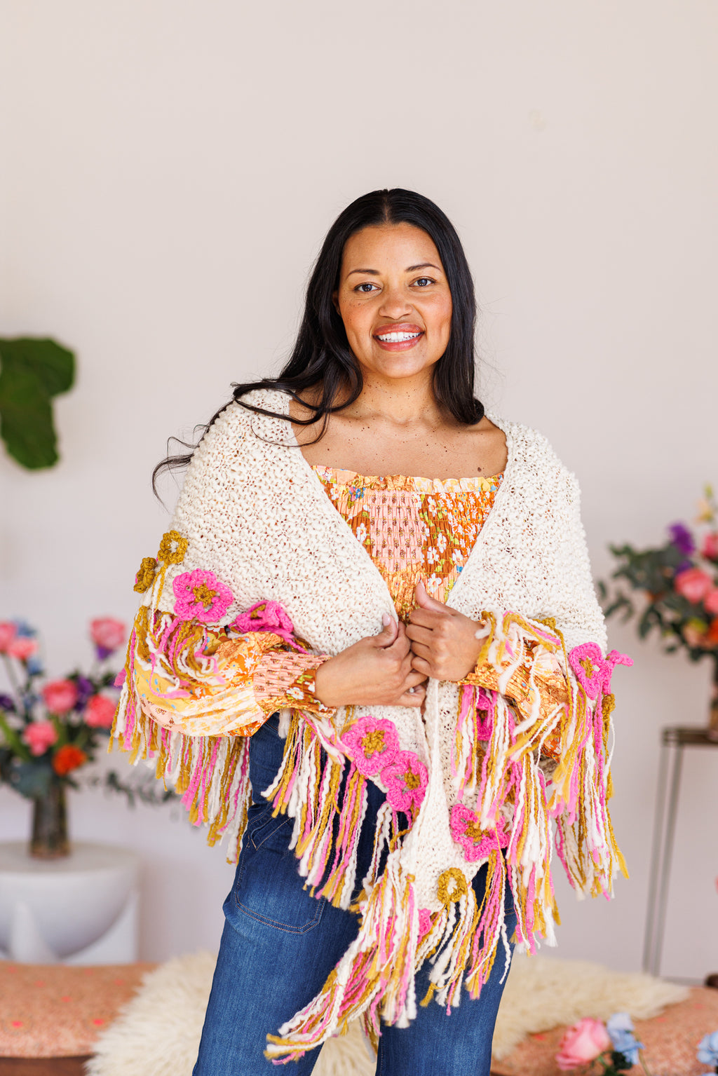 Smiling model holding cream knit shawl closed in front displaying fringe and crochet flower details
