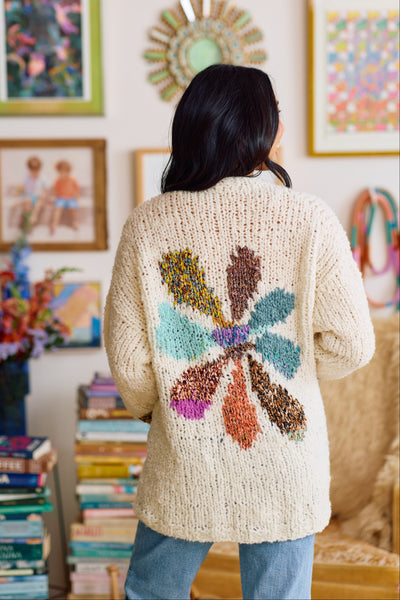 Person wearing a beige hand knit cardigan with a colorful intarsia flower motif on the back, standing in a room with books and artwork.