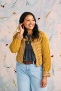 Woman wearing a mustard yellow cardigan over a black top and blue jeans, standing against a decorative wall.