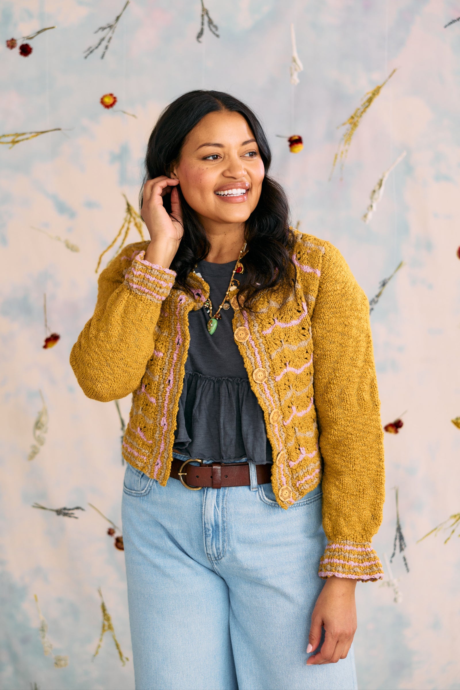 Woman wearing a mustard yellow cardigan over a black top and blue jeans, standing against a decorative wall.