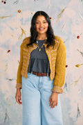 Woman wearing a mustard yellow cardigan over a dark top and light blue jeans, standing against a decorative wall.