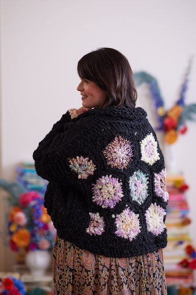 Smiling model displaying back of black knit and crochet cardigan featuring pastel crochet hexagons