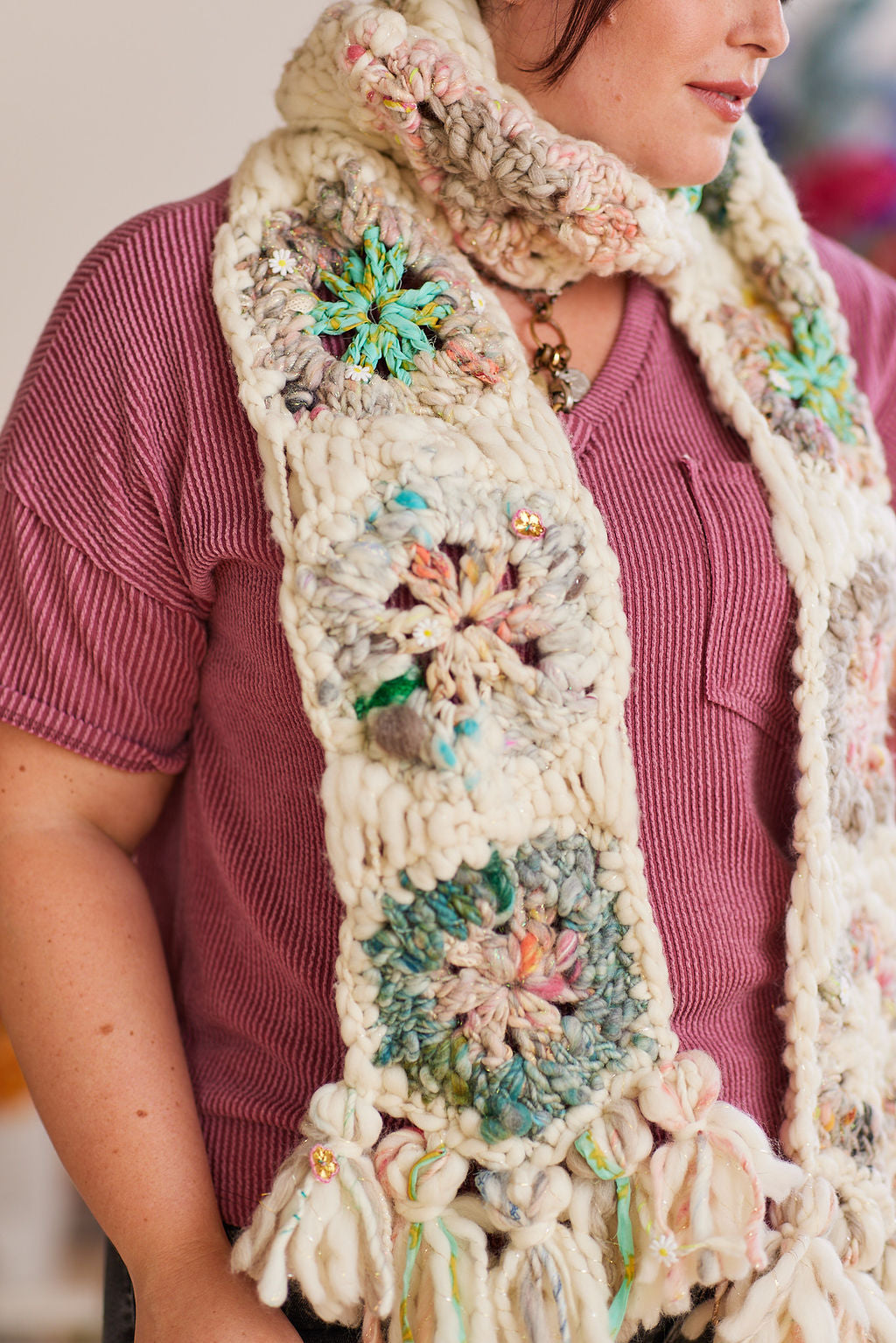 Model is wearing mauve short sleeve shirt with wool scarf displaying several crochet hexagons on one side of scarf including tassels at the end