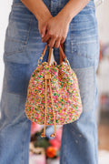 Model holding bucket bag with both hands in front of denim jeans, displaying the closure of the drawstring in front as well as the metal hardware connecting strap to top sides of bag