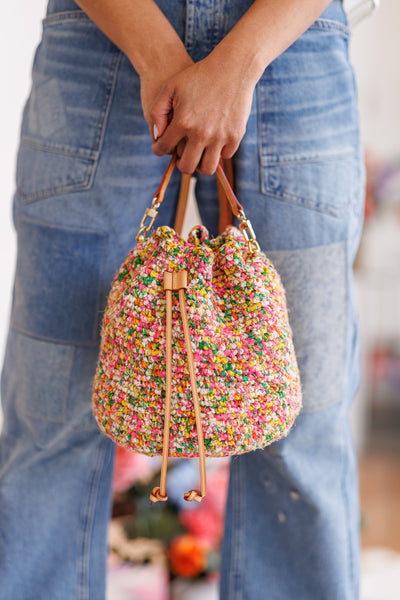 Model holding bucket bag with both hands in front of denim jeans, displaying the closure of the drawstring in front as well as the metal hardware connecting strap to top sides of bag