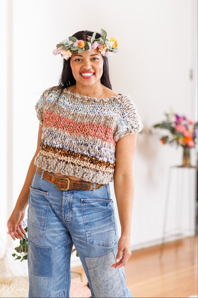 A smiling woman standing in a room with a floral wreath on her head, wearing a multicolored cropped sweater with a striped pattern, paired with blue denim pants.