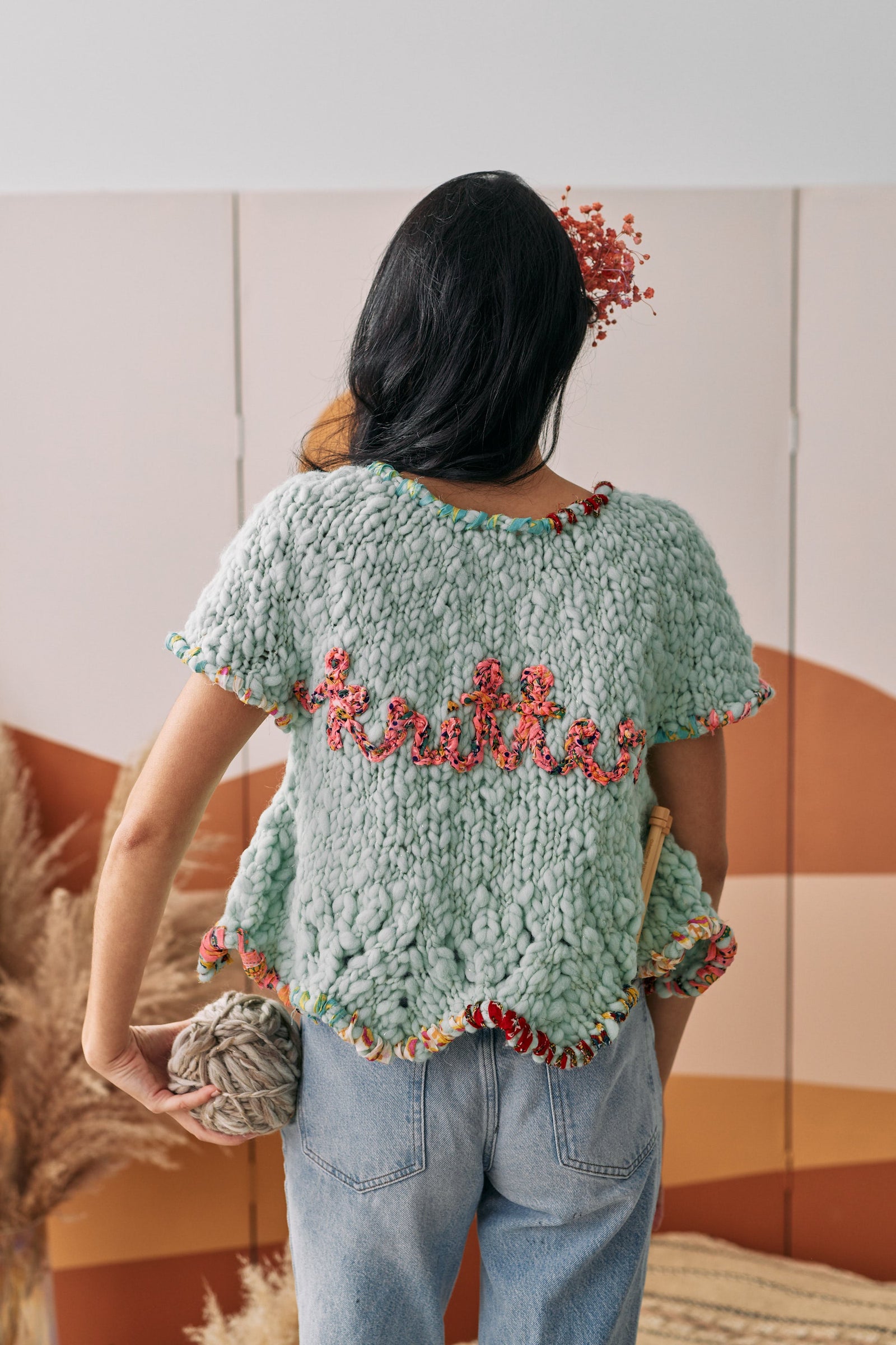 Woman holding yarn with knitting needles in her back pocket, displaying her cardigan that says 'knitter'