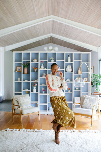Woman standing in front of a bookshelf models a cream colored cardigan over a gold dress