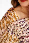 Close-up of yarn and stitch details on knit wrap's edge as it drapes over model's shoulder, wrap held closed with brass shawl pin