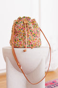 Multicolored, cotton, crochet bucket bag sitting on a pedestal displaying adjustable leather strap, hardware, and brown leather drawstring closure