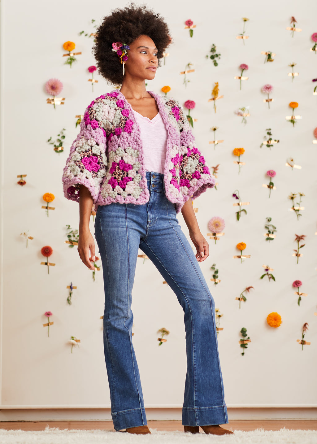 Model posing in front of flower wall wearing lavender and orchid crochet granny square cardi over a V-neck t-shirt with denim jeans and heeled boots