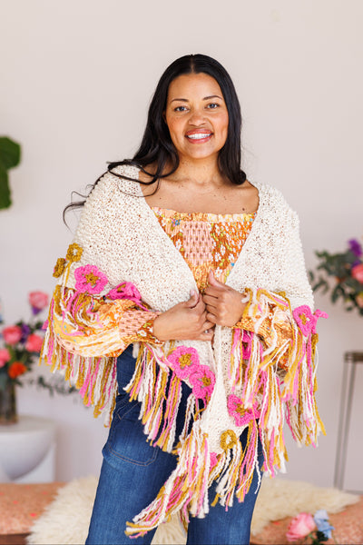 Smiling model holding cream knit shawl closed in front displaying fringe and crochet flower details